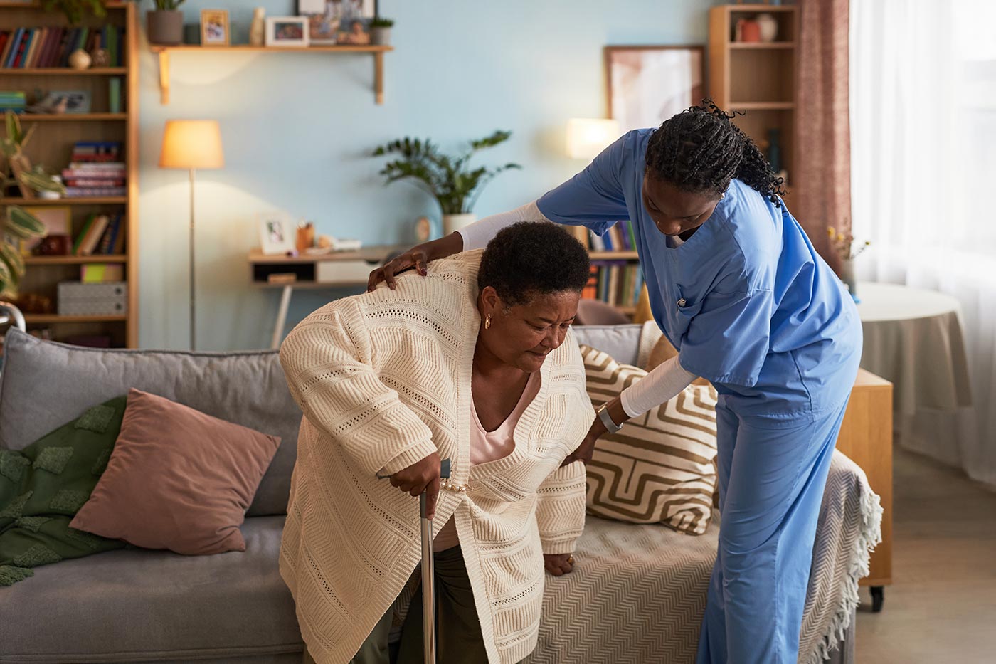 A carer in uniform helping an elderly woman up from her sofa at home
